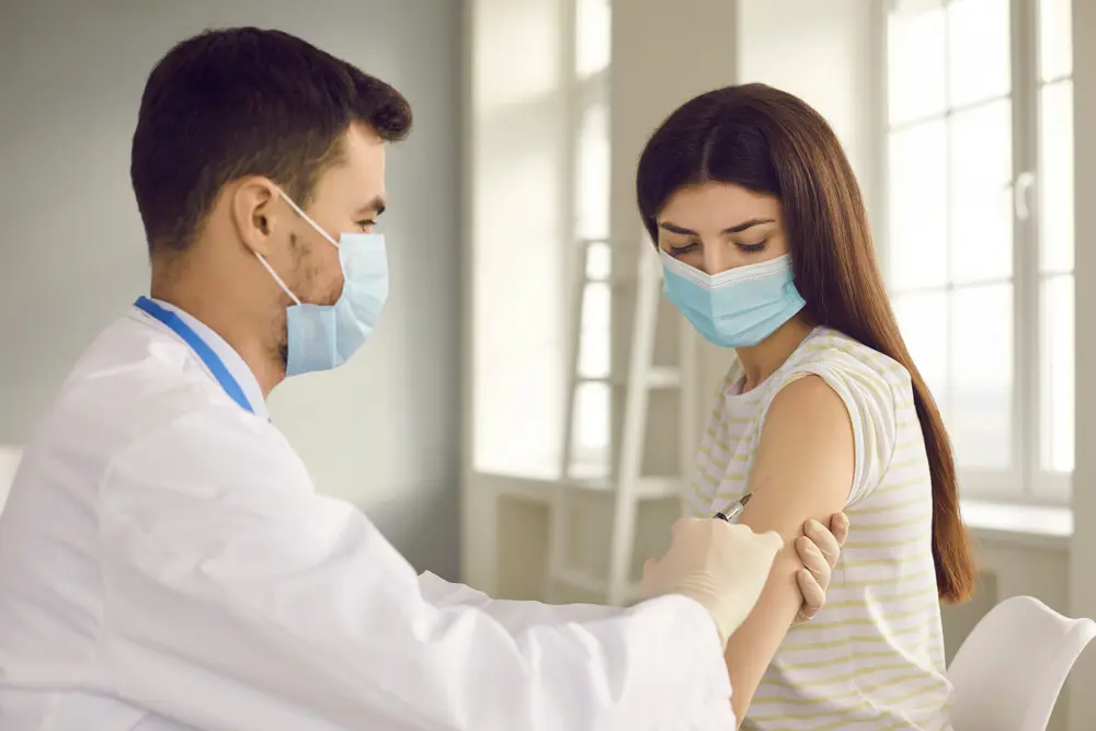 Male nurse or doctor in medical face mask giving flu injection to female patient during seasonal immunization campaign at health center