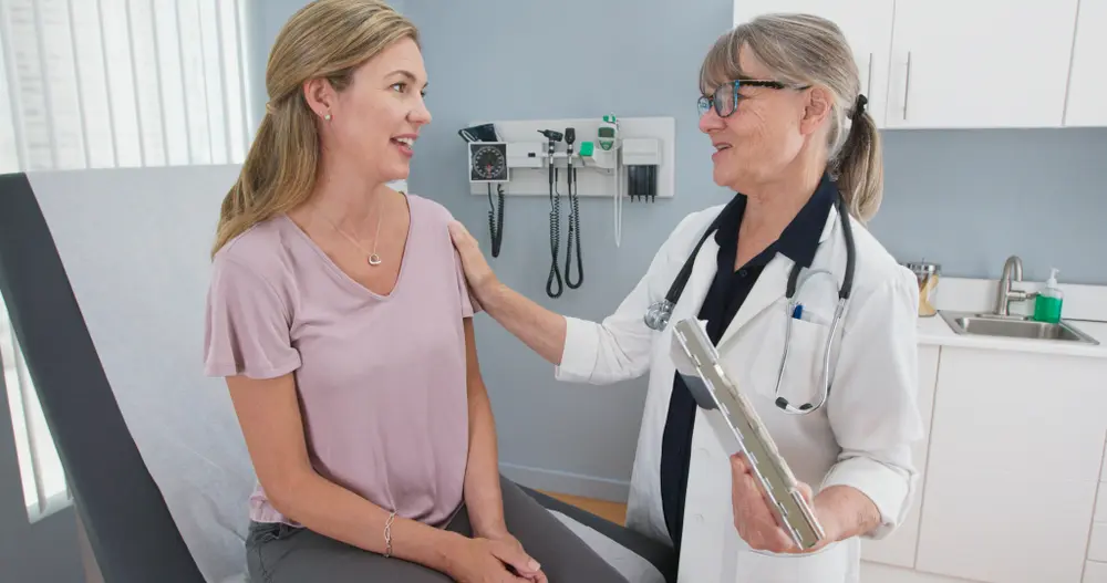 A doctor with a stethoscope speaks kindly to a woman sitting on an exam table in a medical office. The doctor has a clipboard and rests a reassuring hand on the patient’s shoulder. Medical equipment is visible in the background.