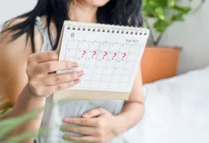 A woman holds a calendar showing August 2021, with several dates marked with red question marks. She is touching her stomach, suggesting concern or uncertainty, with green plants visible in the background.