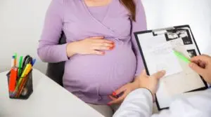 A pregnant woman in a purple top sits at a desk with her hands on her belly while a healthcare professional reviews medical documents on a clipboard, discussing warning signs during pregnancy.
