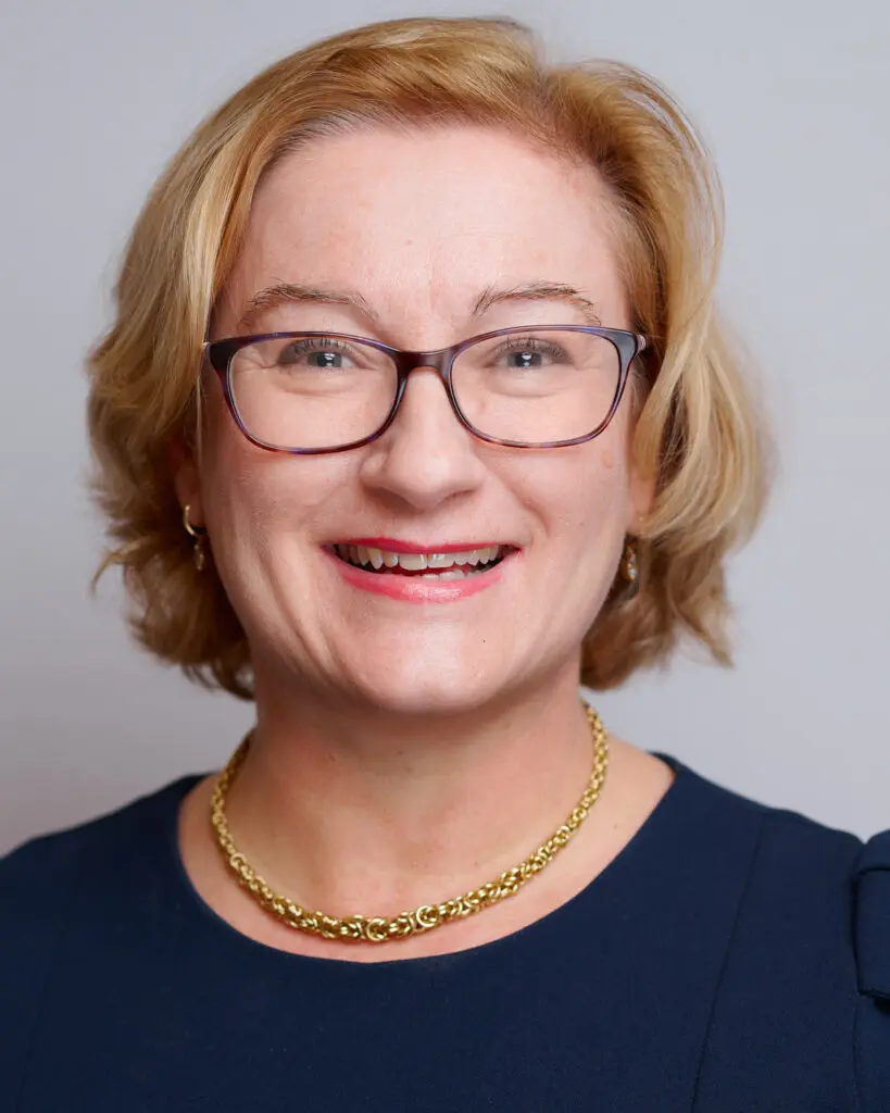 A woman with short blonde hair and glasses is smiling. She is wearing a dark blue top, a gold necklace, and small hoop earrings, and is standing in front of a plain light background.