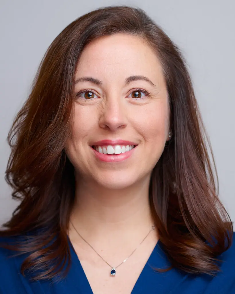 A woman with long brown hair and brown eyes smiles at the camera. She is wearing a blue top and a silver necklace with a dark pendant. The background is plain and light gray.