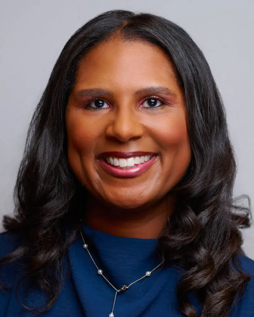 A woman with long, wavy dark hair smiles at the camera. She is wearing a navy blue top and a delicate necklace with small pearls. The background is plain and light-colored.