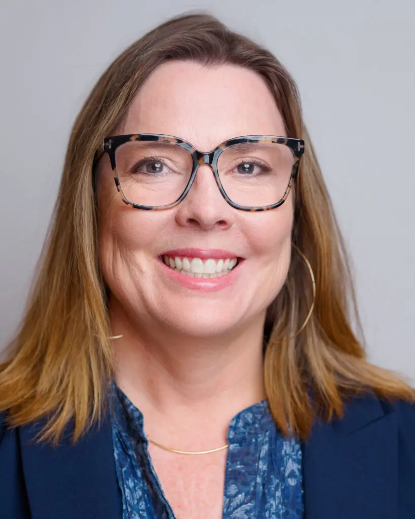 A woman with straight, shoulder-length light brown hair, wearing large tortoiseshell glasses, a blue patterned blouse, a dark blazer, and a thin gold necklace, smiling at the camera against a neutral background.