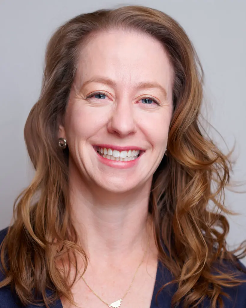A woman with wavy, light brown hair smiles at the camera. She is wearing a dark top, a necklace, and small earrings against a plain, light background.