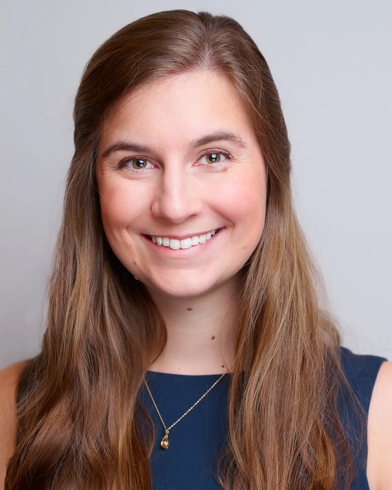 A woman with long brown hair and a navy blue sleeveless top smiles at the camera. She is wearing a gold necklace and standing in front of a plain light gray background.