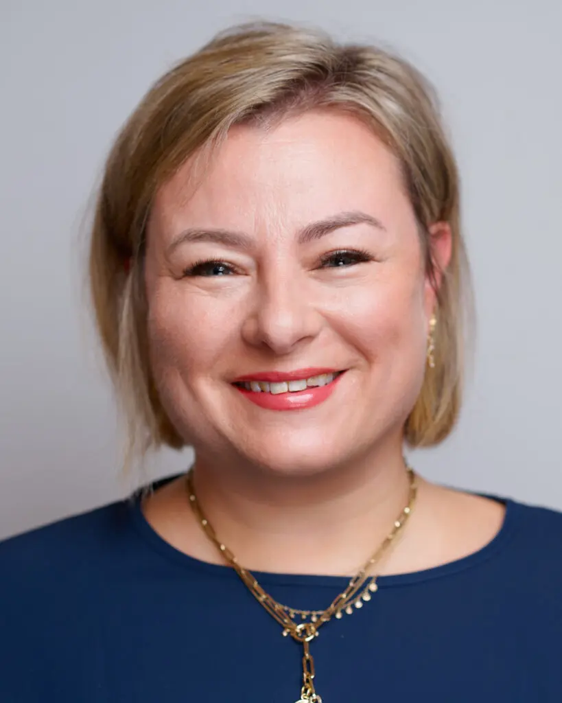 A woman with short blonde hair smiles at the camera. She is wearing a navy blue top, gold earrings, and layered gold necklaces, with light makeup and a neutral background.