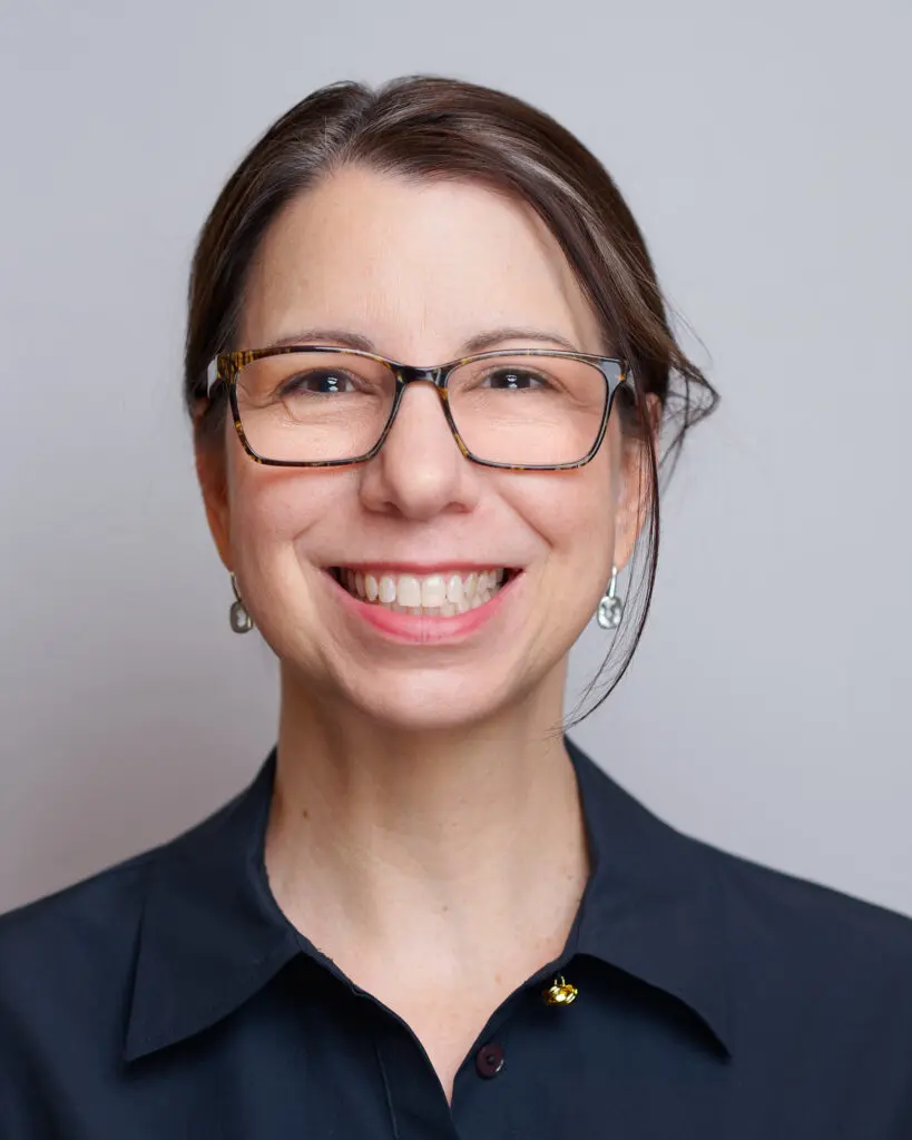 A woman with brown hair in a low bun, wearing glasses, silver earrings, and a dark collared shirt, smiles at the camera against a plain light gray background.