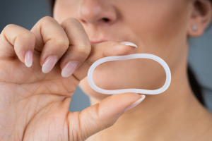 A person holding a flexible, oval-shaped contraceptive vaginal ring between their fingers, with their face partially visible in the background.