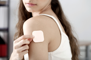 A person wearing a white tank top applies a beige adhesive patch to their upper arm, suggesting the use of a medical or nicotine patch. The background is blurred.