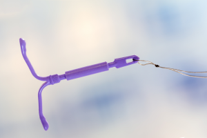 A close-up of a purple intrauterine device (IUD) with attached strings, used for long-term birth control, against a blurred light background.