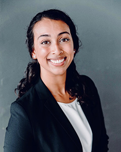A woman with long dark hair smiles at the camera, wearing a black blazer over a white top, standing in front of a plain gray background.